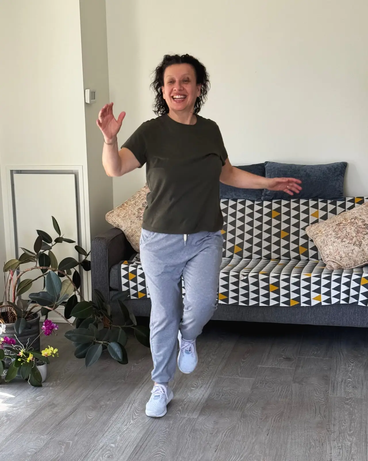Diana dancing in her living room in front of the sofa, mid-step with both arms open