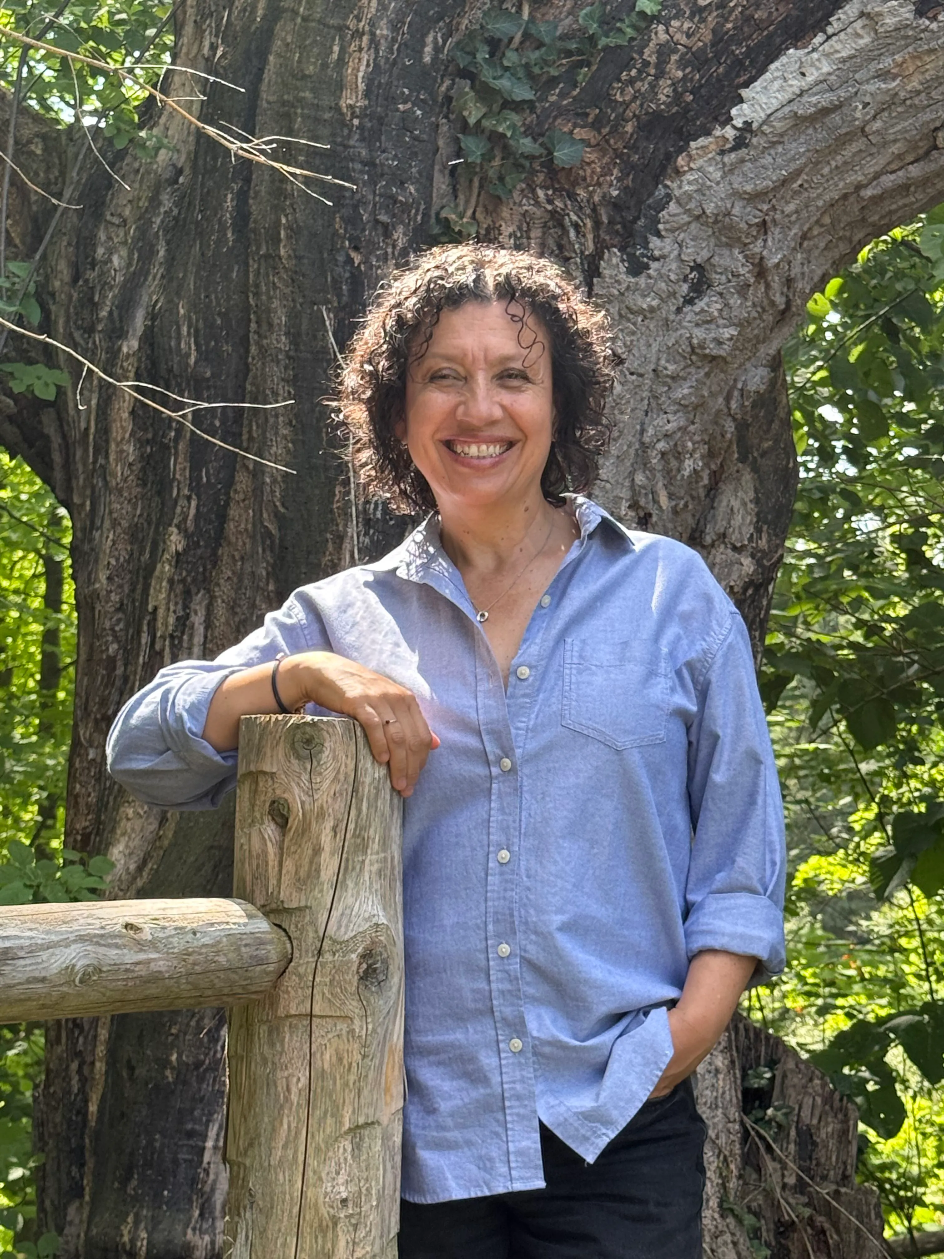 Diana smiling, leaning on a cedar fence post in front of a large old tree