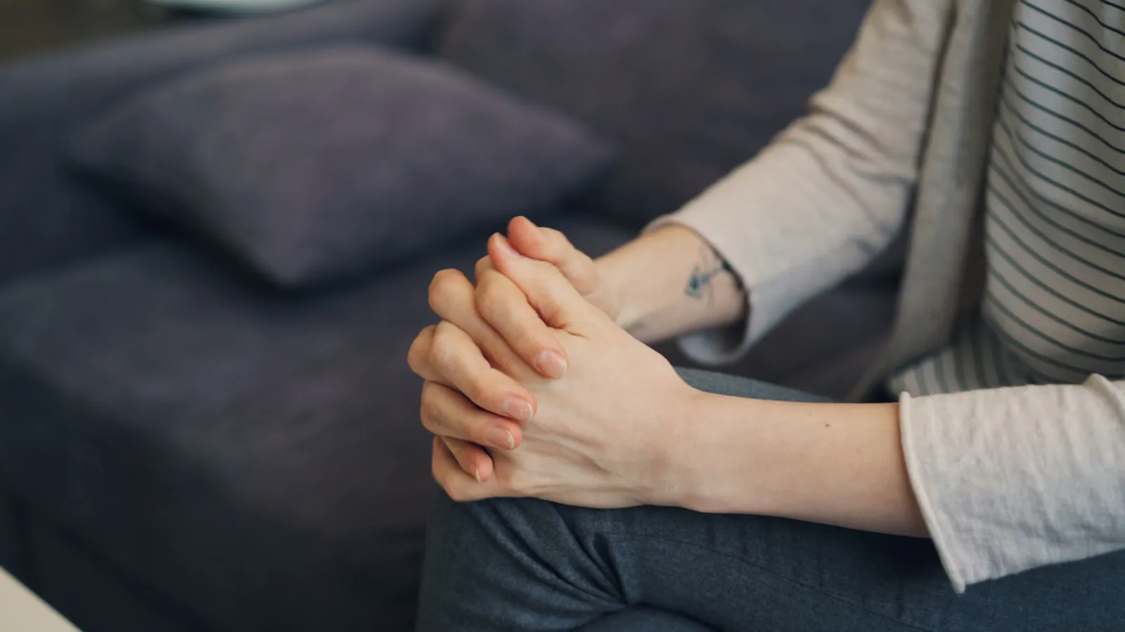 A person seated on a sofa, hands clasped loosely in their lap