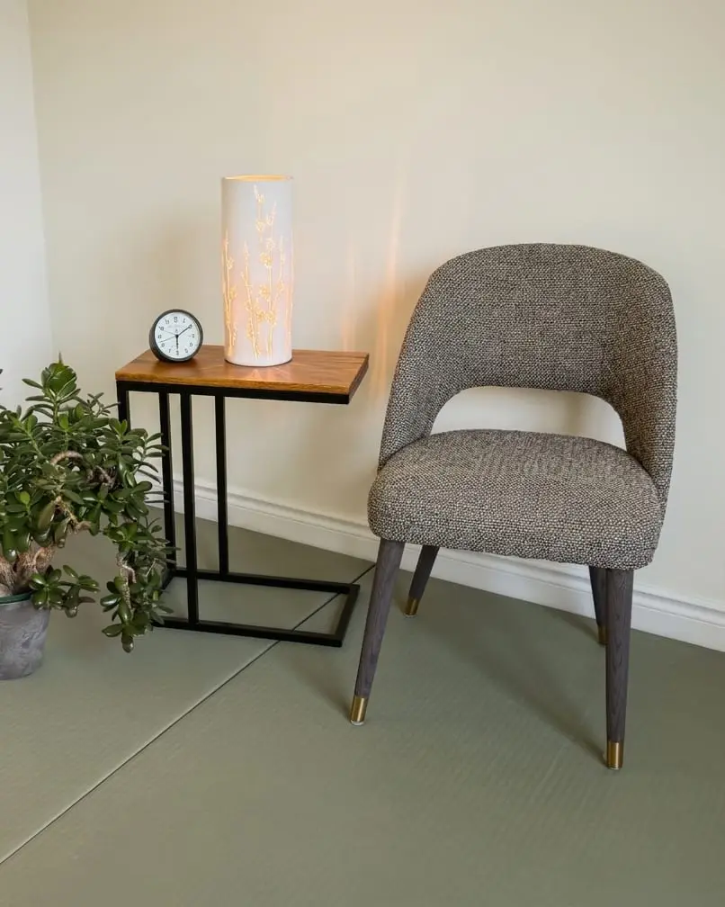 A corner of Diana's room: a soft grey armchair on a tatami floor, beside a small wooden table with a lit lamp and a clock, and a jade plant